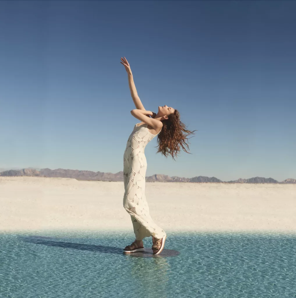 A woman standing in a shallow body of water in a desert landscape, reaching her arm upwards with her face turned upward.