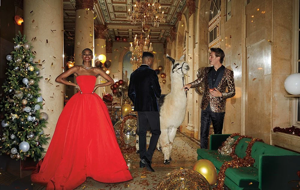 People celebrating Christmas in an ornate, decorated hall with a Christmas tree, balloons, and confetti. One woman in a red gown, two men interact with a llama, and festive decorations are everywhere.