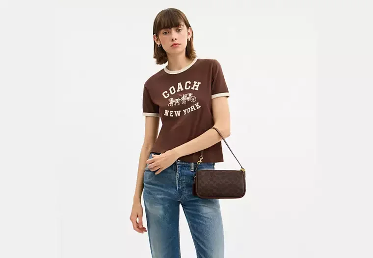 Young woman with brown hair wearing a brown T-shirt with white lettering and design, standing against a plain background, holding a small dark designer handbag.
