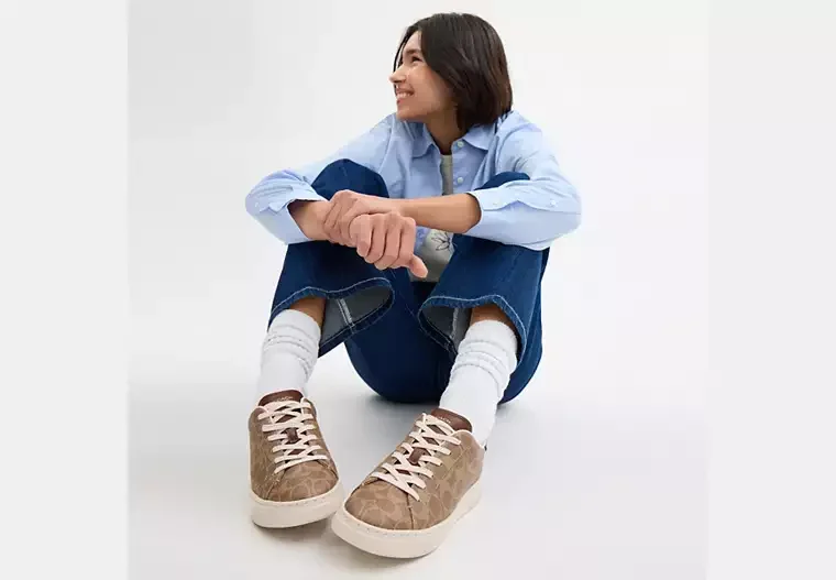 Woman sitting on the floor against a white background, smiling and looking to the side, dressed casually in a blue shirt, blue jeans, white socks, and beige sneakers.