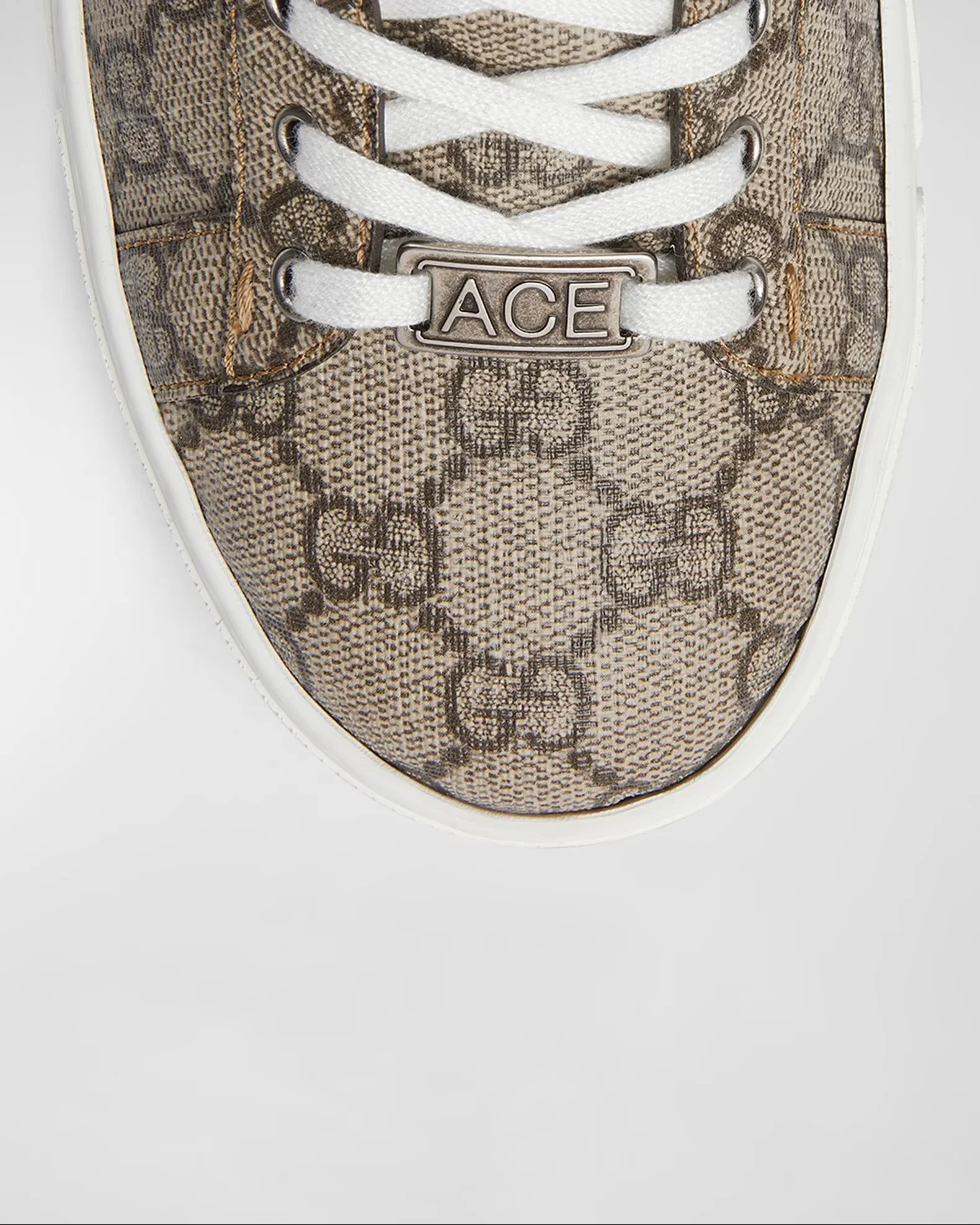 Close-up of a beige and brown designer sneaker with white laces and metal eyelets, featuring a metal tag engraved with the name 'ACE' on the tongue.