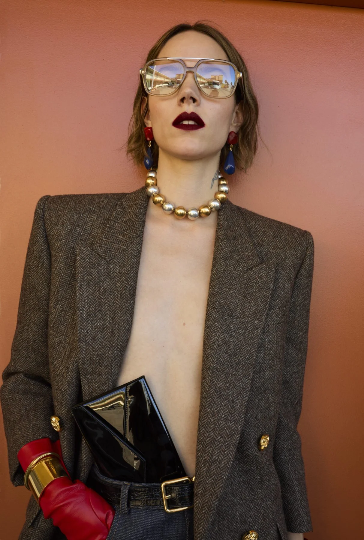 Fashionably dressed woman wearing large reflective glasses, bold lipstick, red and blue earrings, a pearl necklace, a brown blazer, holding a black clutch, and wearing a red glove with gold accents, standing against a pink wall.