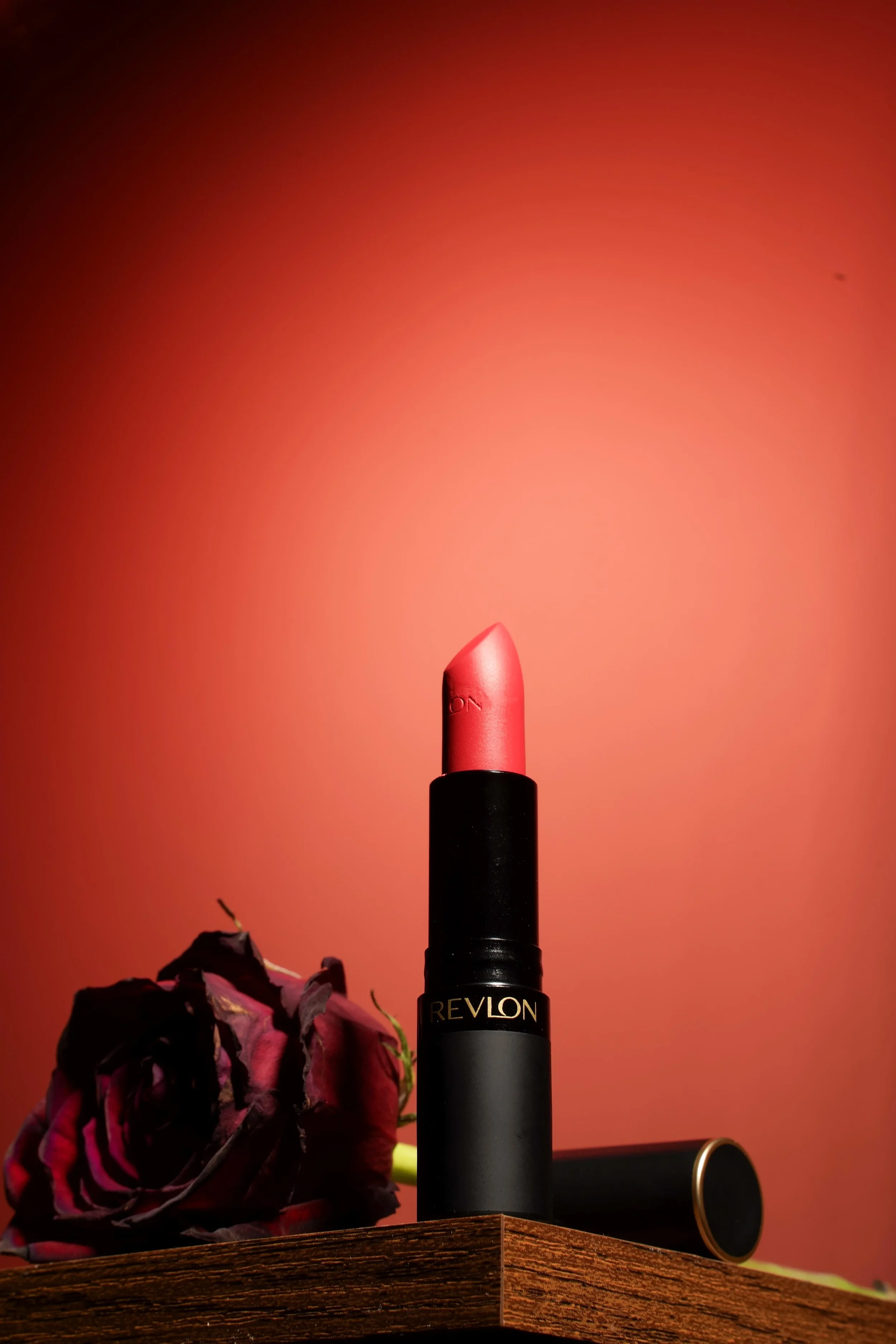 A Revlon lipstick in a black tube with the red lipstick extended, placed on a wooden surface next to a dried, dark pink rose and a black makeup container with a gold rim, against a warm orange background.