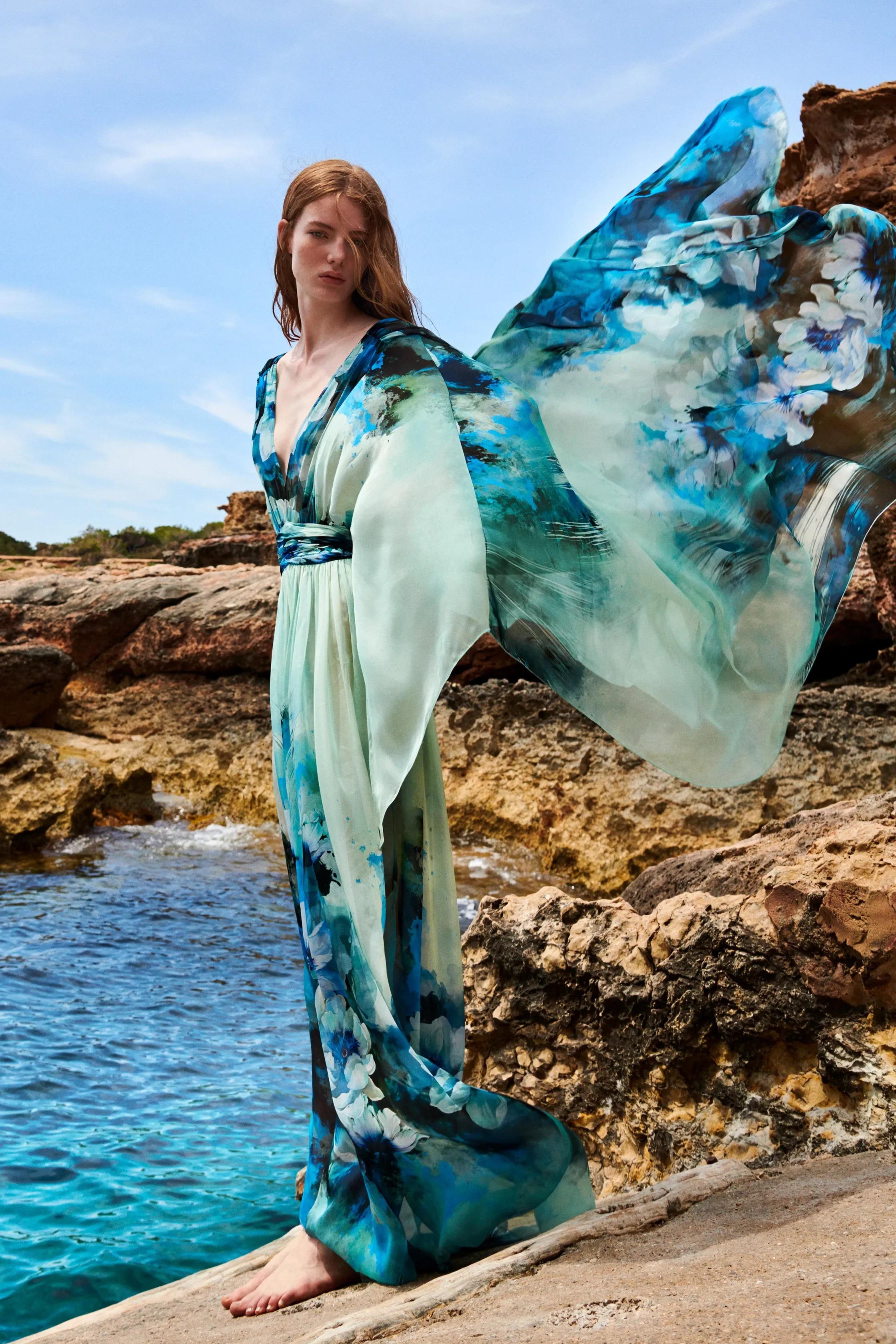 A young woman with red hair standing on rocks by the ocean, wearing a flowing, blue and green floral dress, with the wind blowing her dress and hair.