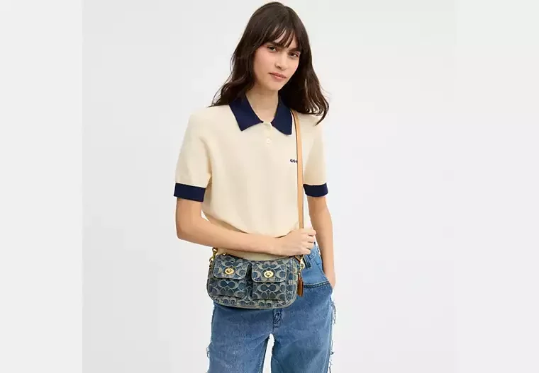 Young woman with dark brown hair wearing a cream and navy polo shirt, blue jeans, and carrying a Louis Vuitton crossbody bag, standing against a plain white background.