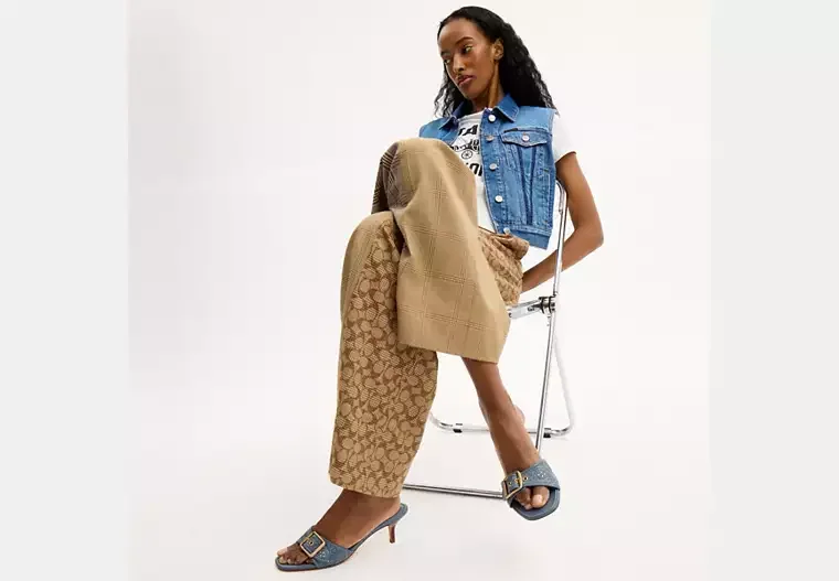 Woman in trendy clothing sitting on a clear chair against a white background, wearing a blue sleeveless denim vest, beige patterned pants, and navy high-heeled sandals.