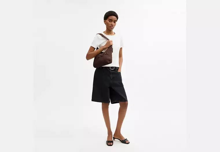 A woman standing against a plain white background, wearing a white T-shirt, black shorts, black sandals, and holding a brown textured shoulder bag.