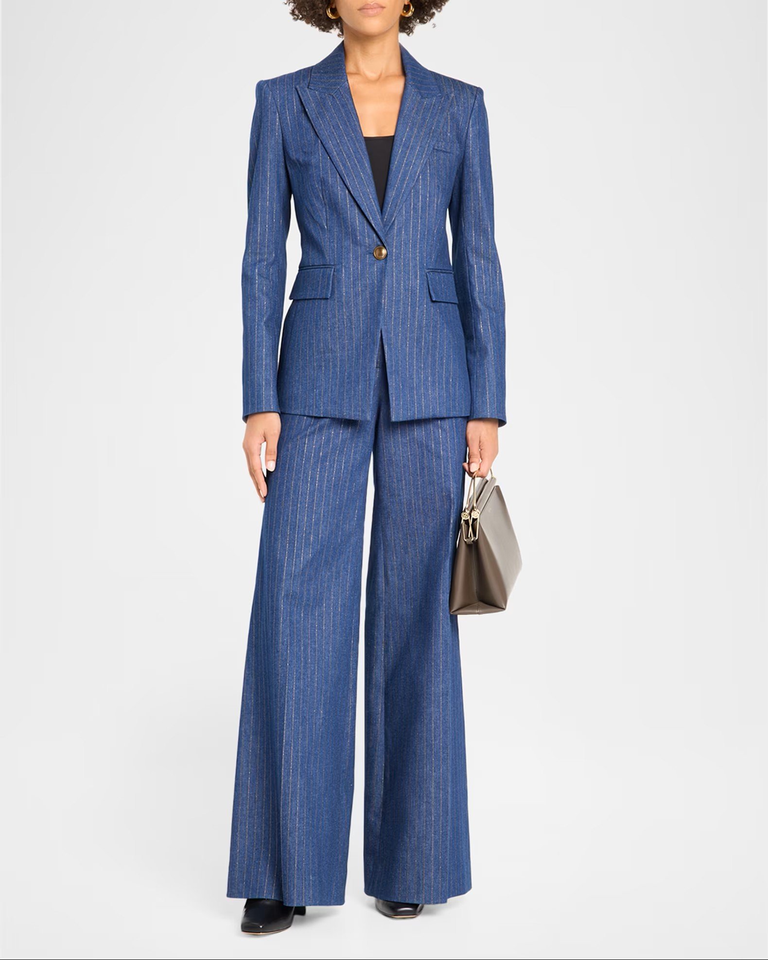 A woman wearing a blue pinstripe blazer and matching wide-leg pants, holding a beige handbag, standing against a plain light background.
