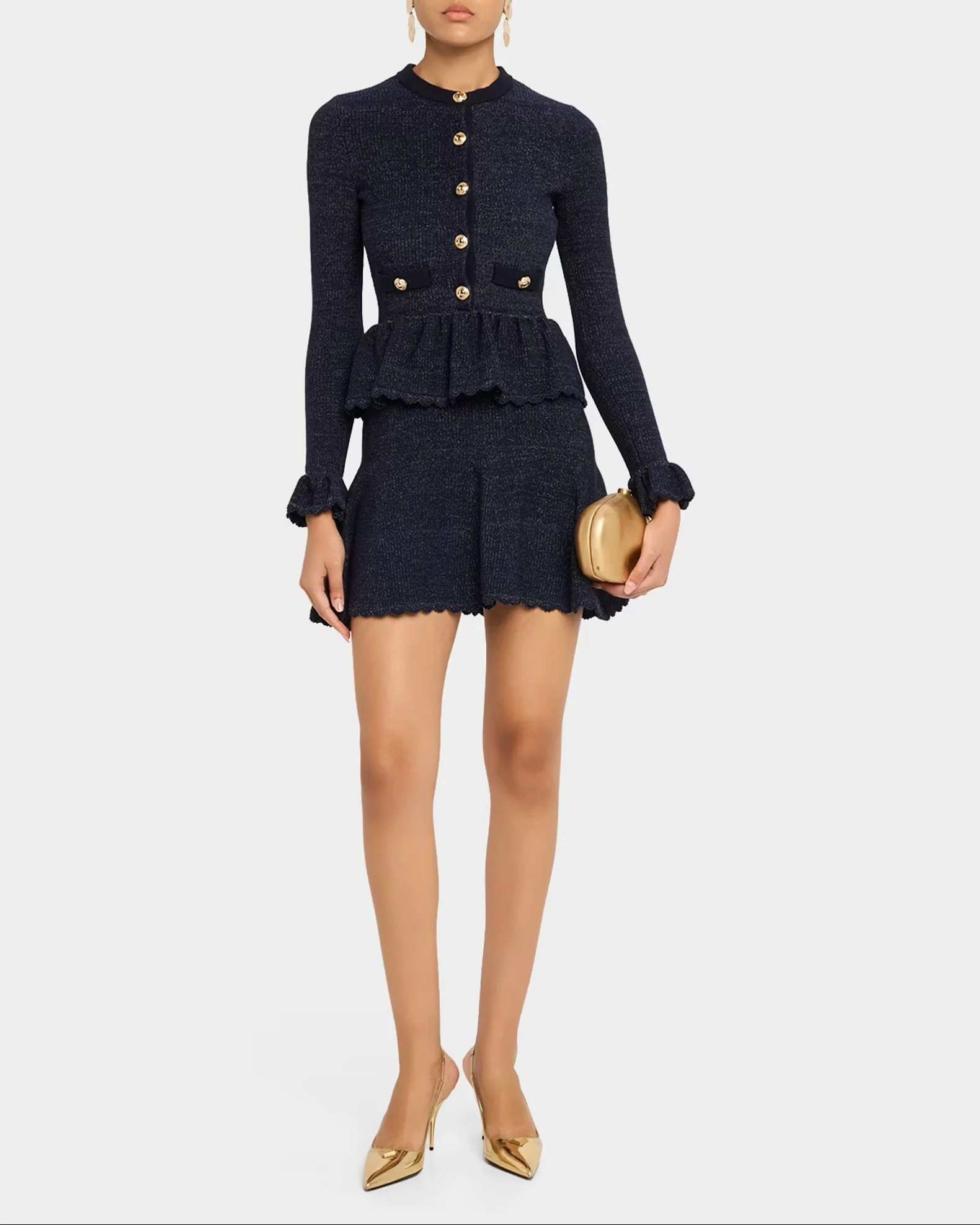 A woman wearing a dark textured long-sleeve dress with gold buttons, ruffled cuffs, and a peplum waist, holding a gold clutch purse, wearing nude high heels and gold earrings.