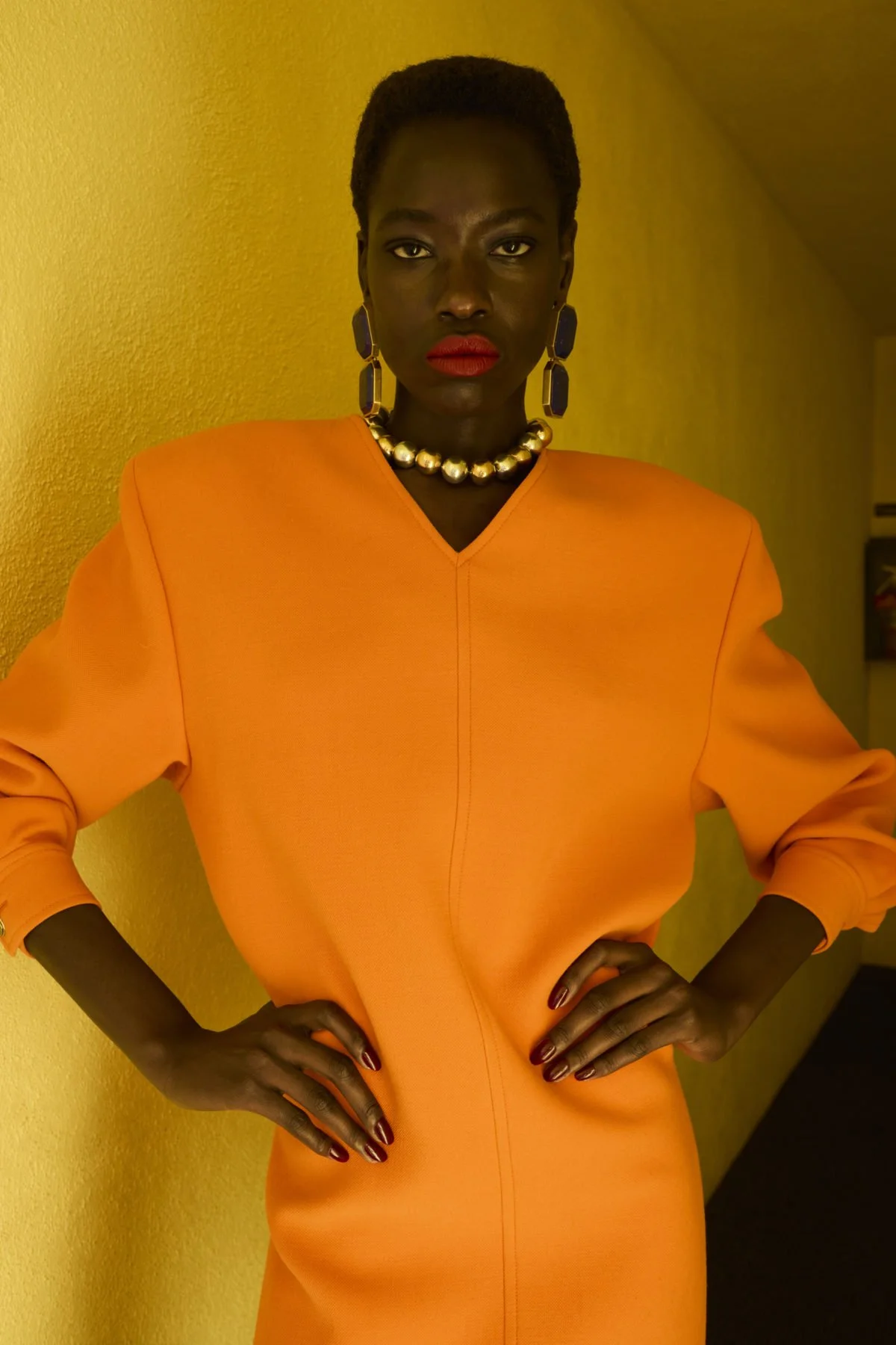 A woman with dark skin wearing an orange dress, large navy blue earrings, a pearl necklace, and red lipstick, standing against a yellow textured wall