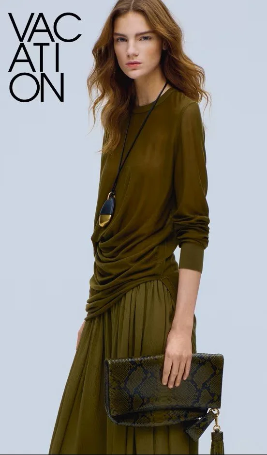 A young woman with long, wavy hair in an olive green outfit holding a snakeskin-patterned clutch, standing against a light gray background with the word 'VACATION' in bold black letters.