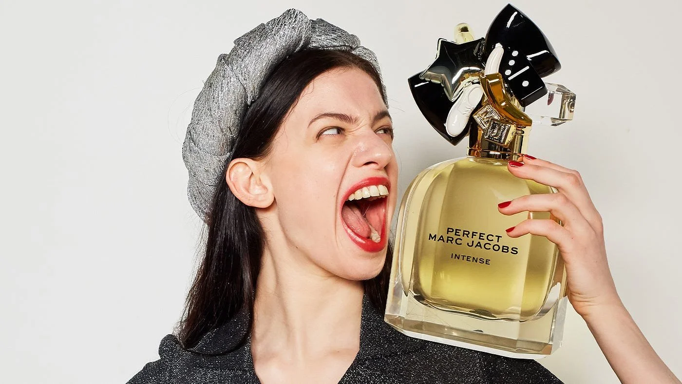 A woman with dark hair, wearing a metallic gray hat and dark sparkly top, holding a large bottle of Marc Jacobs perfume close to her face, appearing to shout or scream at the perfume.
