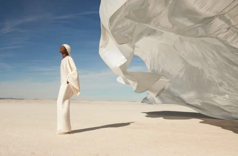 A woman in white standing on a dry, flat landscape with a large white fabric billowing in the wind nearby, under a blue sky with wispy clouds.