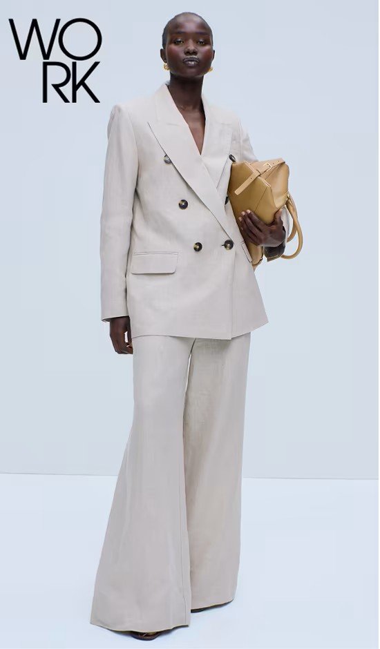 A woman dressed in a beige oversized blazer with matching wide-leg trousers, holding a tan handbag, posing against a plain background with 'WORK' written in black text.