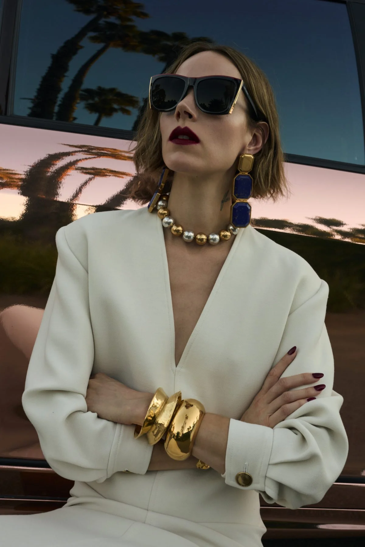 A woman with short bob hair wearing black sunglasses, a white blazer, and gold jewelry, standing in front of a reflective surface showing palm trees at sunset.