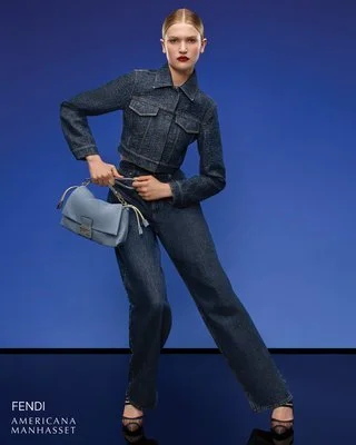 A woman in a denim jacket and jeans posing against a blue background, holding a light blue handbag.