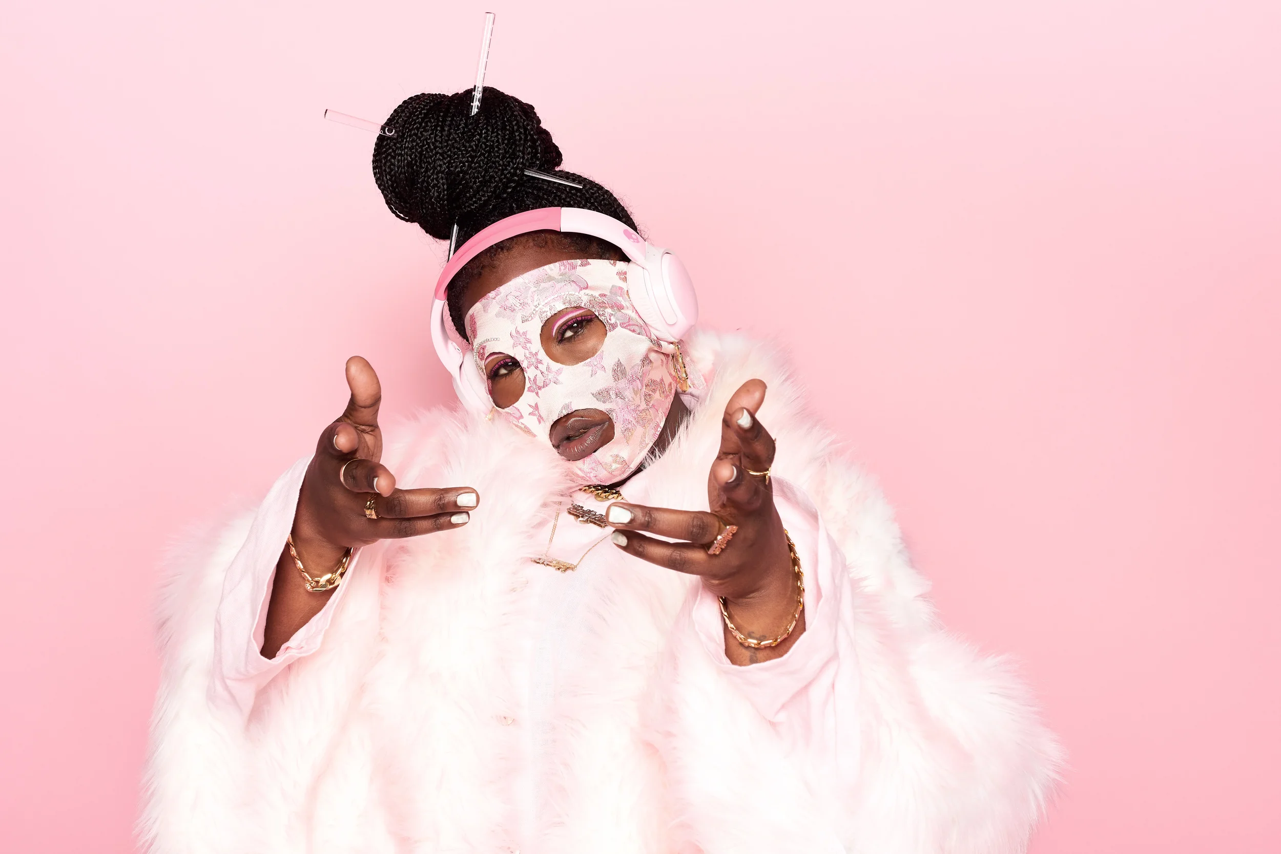 Person with braided hair in a bun, wearing pink headphones and a pink mask with floral patterns, pink background, furry pink coat, jewelry, pointing at the camera.