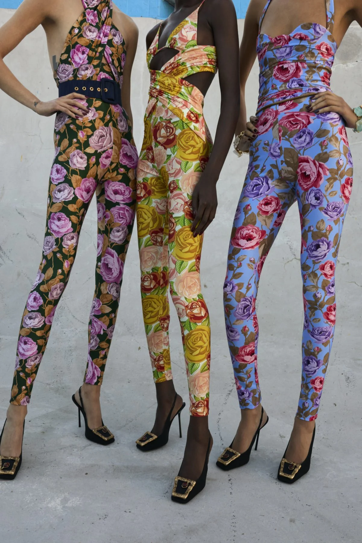 Three women wearing floral patterned jumpsuits and high heels, standing side by side against a plain wall.