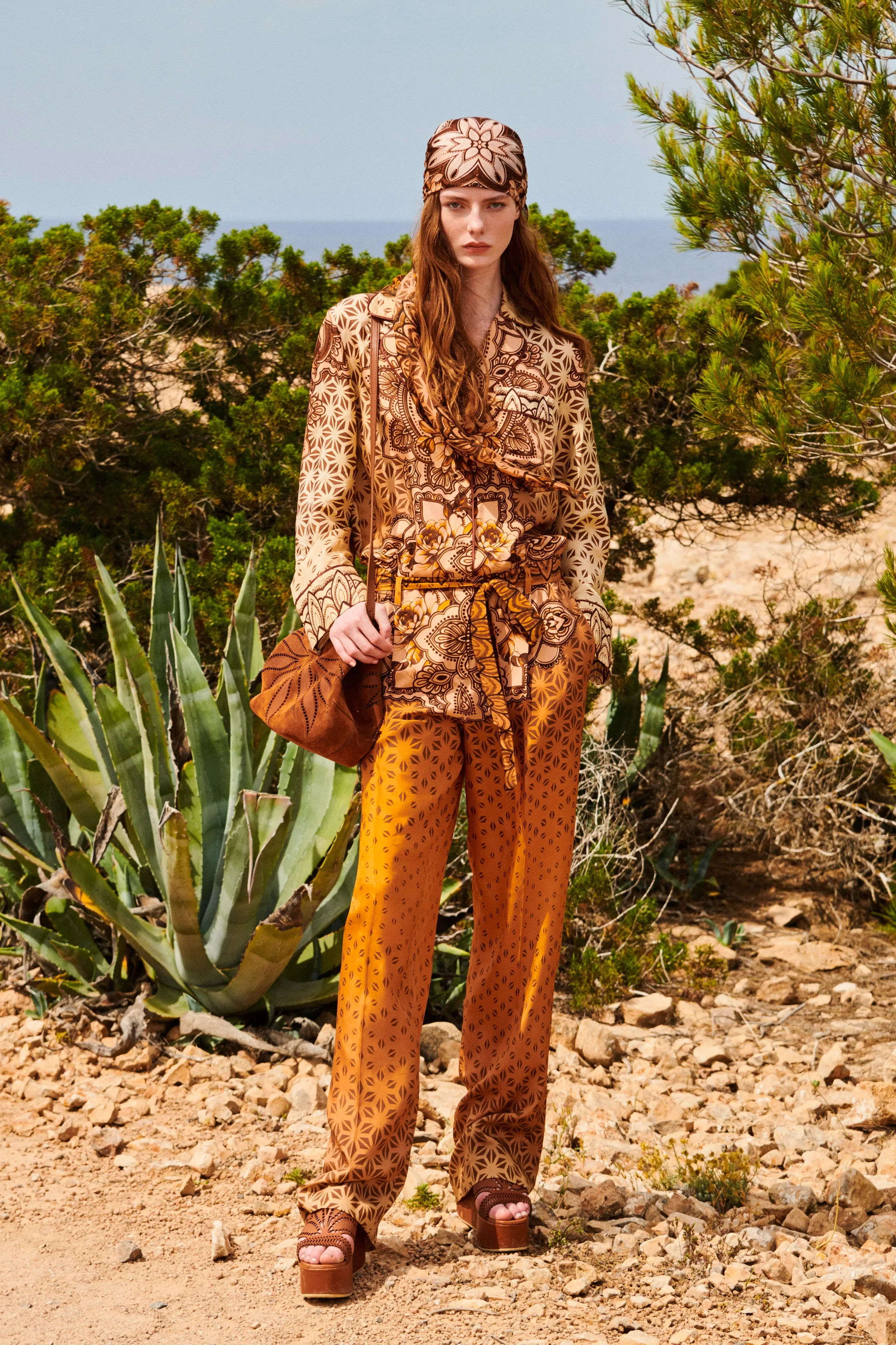 A woman with long red hair standing outdoors among desert plants, wearing an orange patterned blouse, solid orange wide-leg pants, a headscarf, and platform sandals.