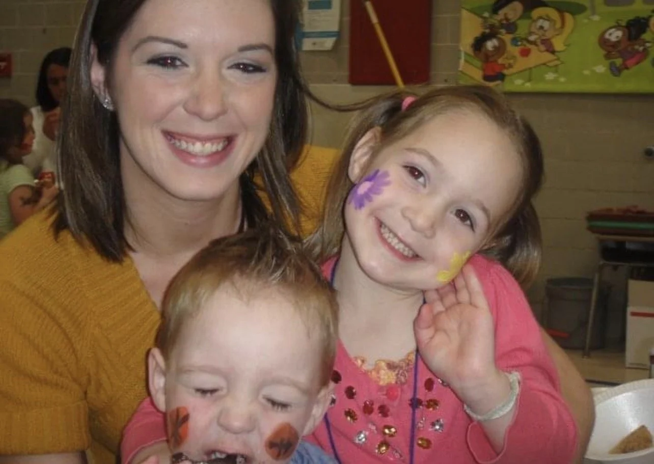 A smiling woman with dark hair and wearing a mustard yellow sweater, holding two young children with face paint in a classroom or indoor setting.