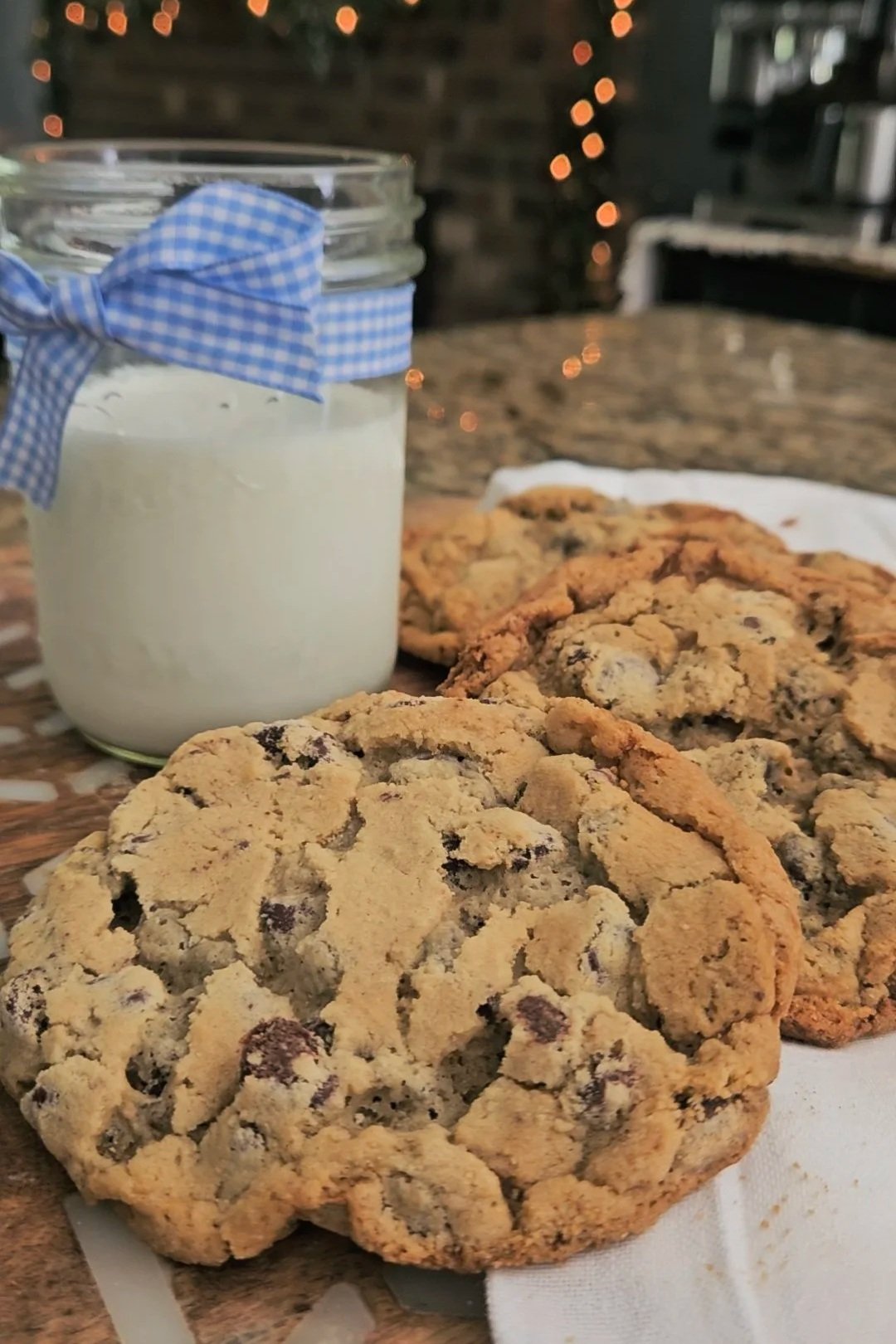 Close-up of chocolate chip cookies and a jar of milk with a blue checkered ribbon tied around its neck, placed on a table with a blurred background of a cozy kitchen decorated with string lights.