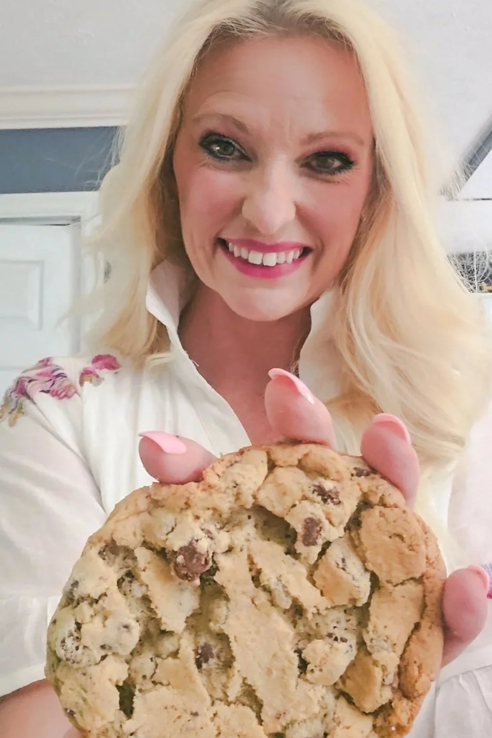 A woman with long blonde hair, smiling and holding a large chocolate chip cookie.