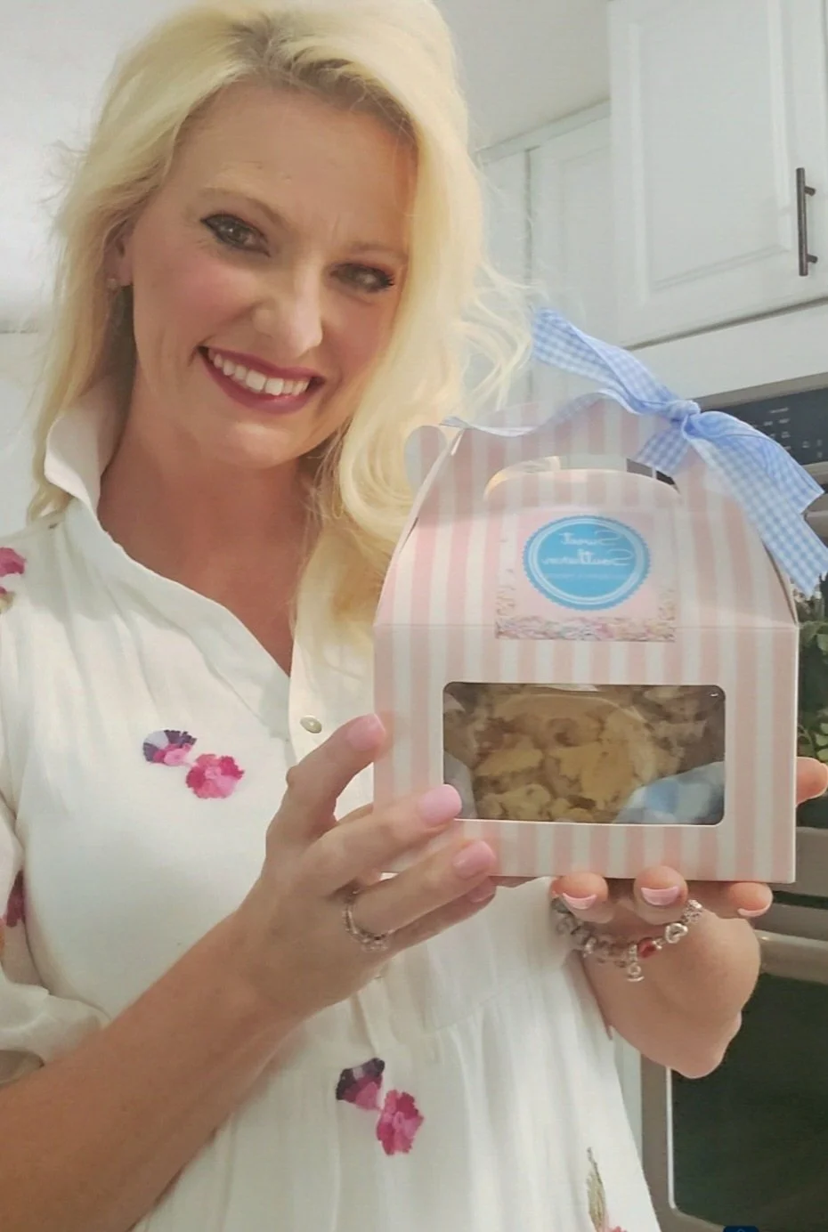 A woman with blonde hair and a floral dress is smiling while holding a pink and white striped box with a clear window showing cookies inside, in a kitchen with white cabinets.