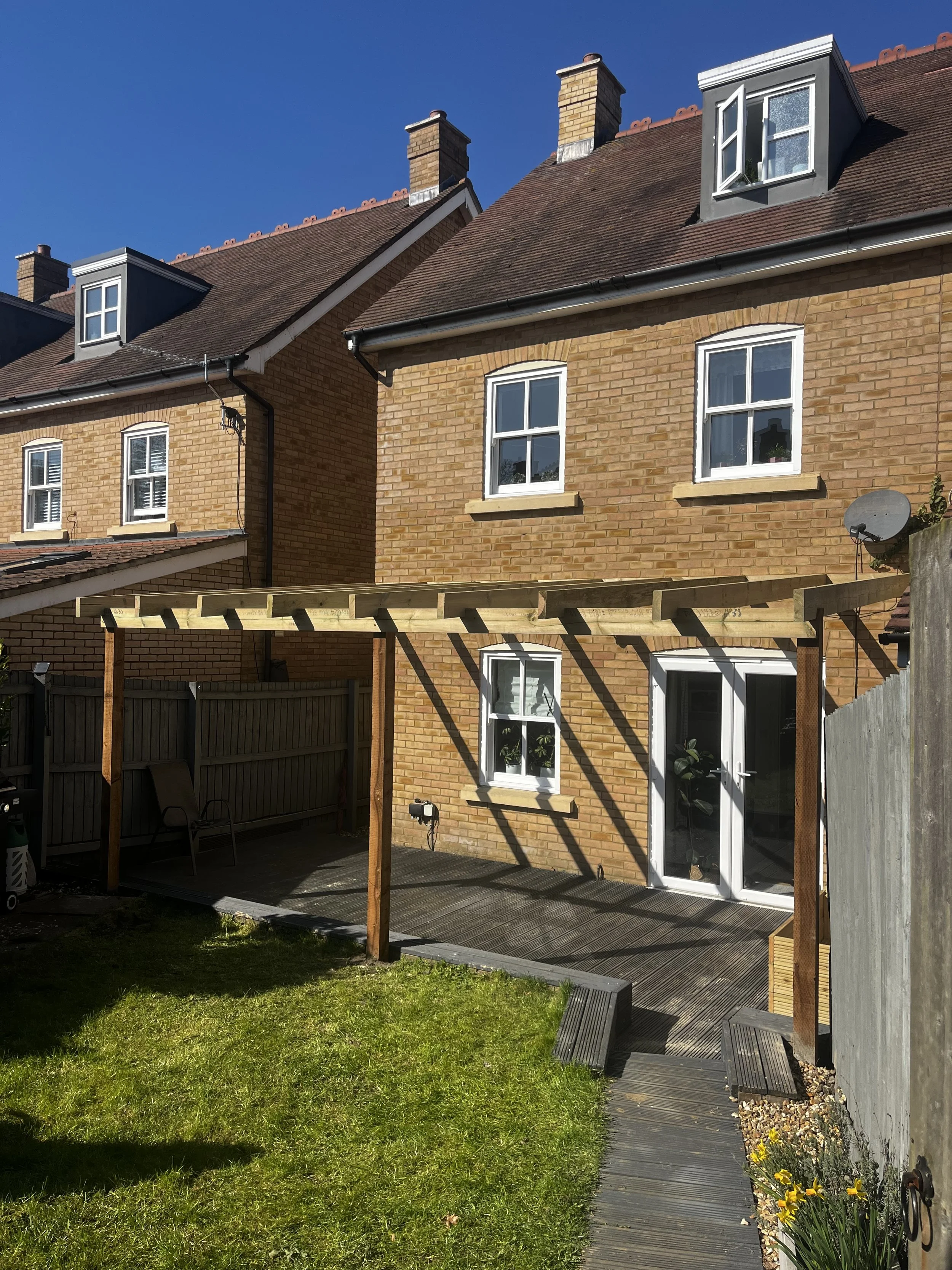 Backyard of a brick house with a wooden pergola, green lawn, and flower bed with yellow flowers, clear blue sky.