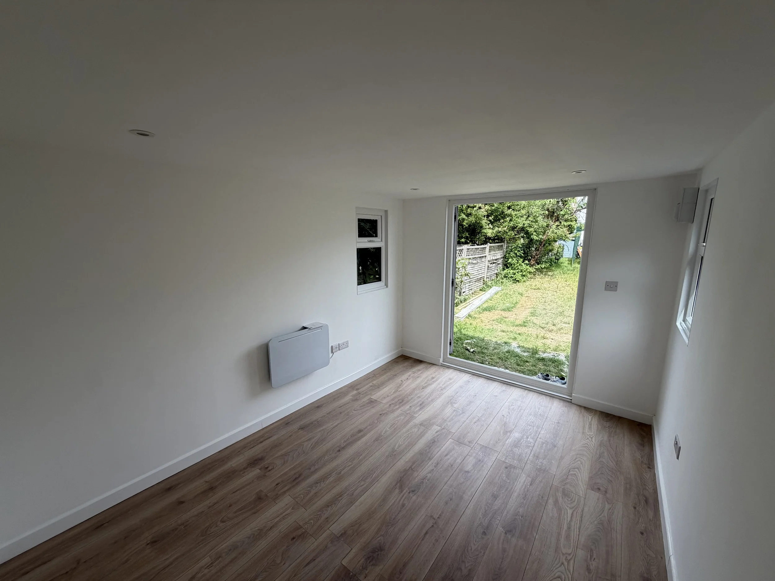 Empty room with white walls, a large open door leading outside to a green yard, two small windows, and wood laminate flooring.