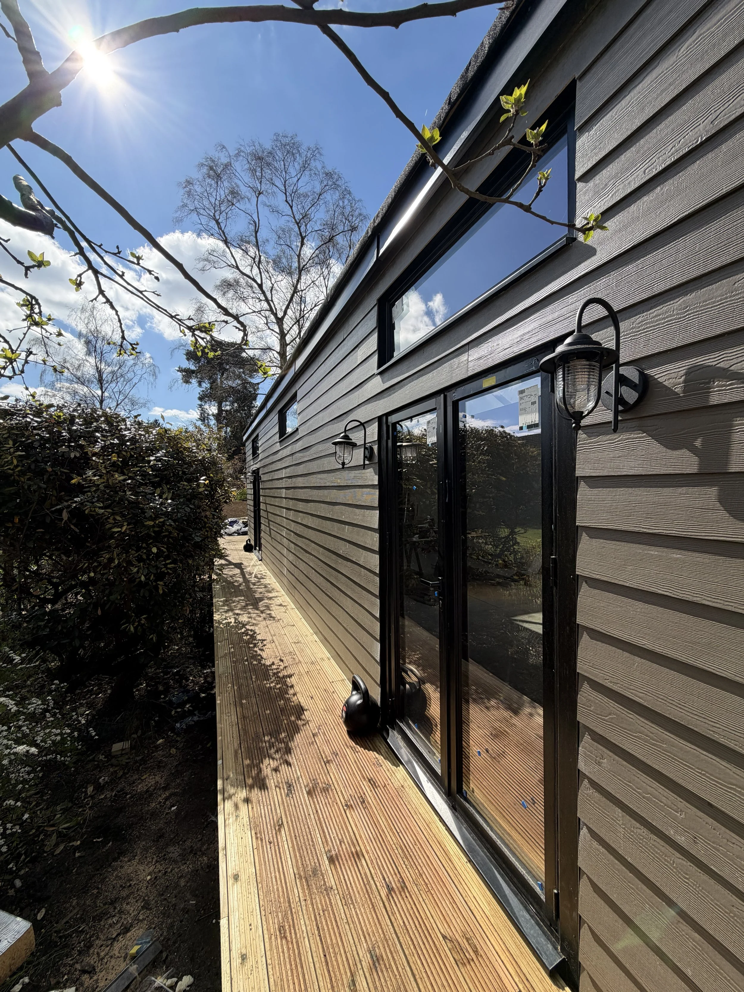 A house with beige horizontal siding, black-framed glass doors, and outdoor wall lights. There is a wooden deck extending from the house, with a small patch of soil and a hedge on one side. Branches with budding green leaves reach toward the sun in a bright, clear sky.