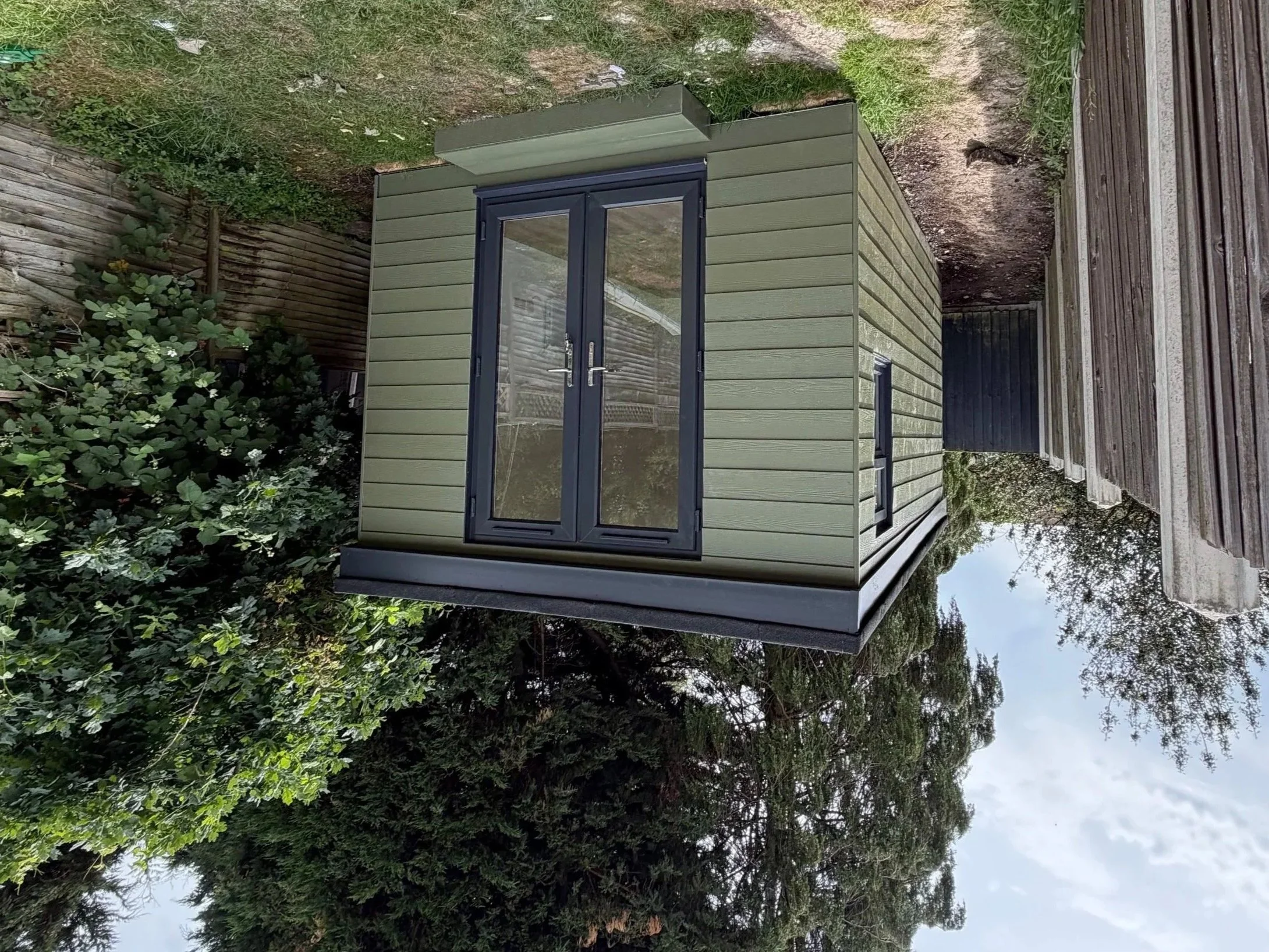 A small green shed with black framed glass doors, located in a backyard with trees and a wooden fence.