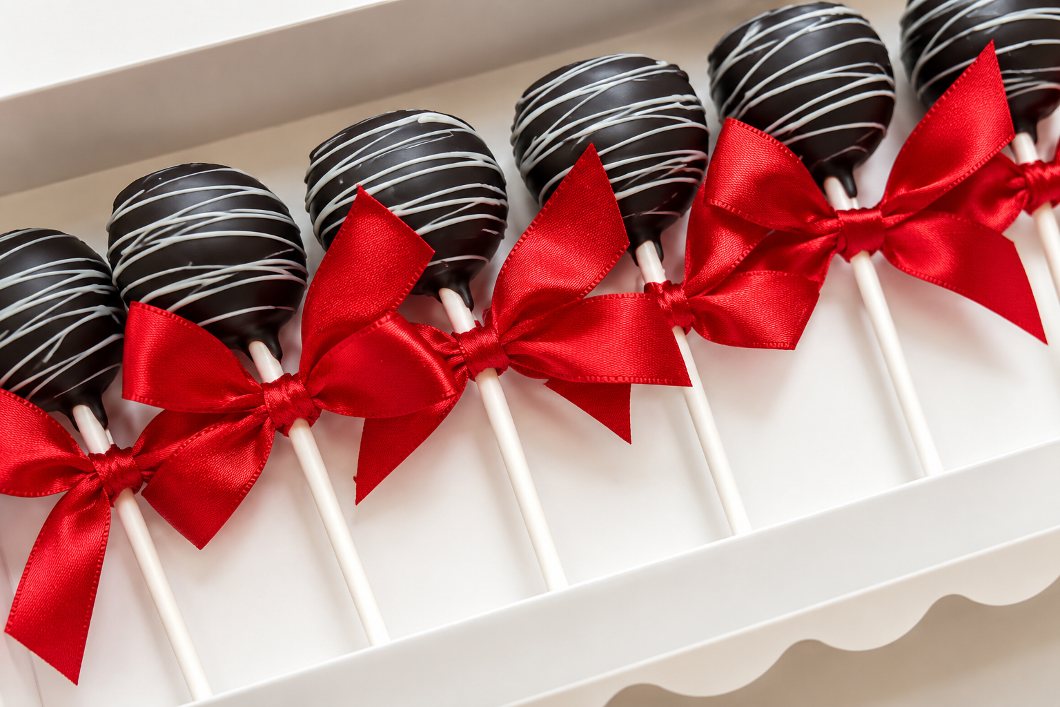 Chocolate cake pops decorated with white drizzle and each tied with a red satin bow, arranged in a white box.