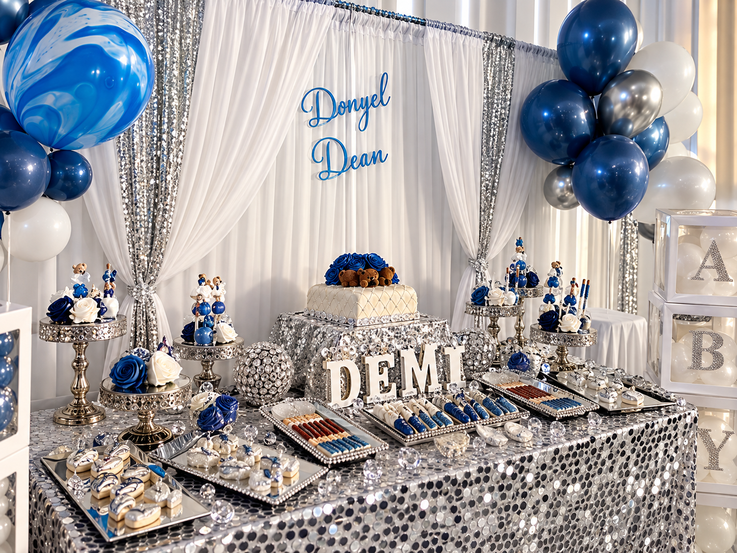 A party table decorated with blue, white, and silver colors, featuring balloons, a cake with teddy bears, and various treats with balloons and a curtain backdrop.