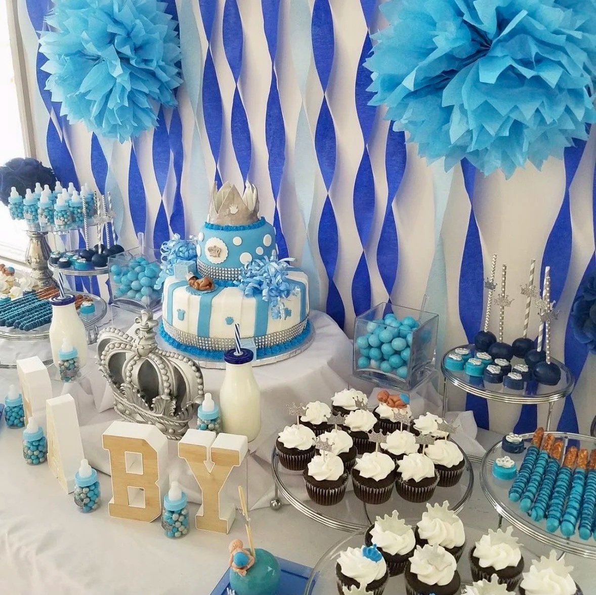 Dessert table at a baby shower with blue and white decorations, cupcakes with white frosting, a blue cake with polka dots and a crown decoration, and blue tissue paper pom-poms in the background.