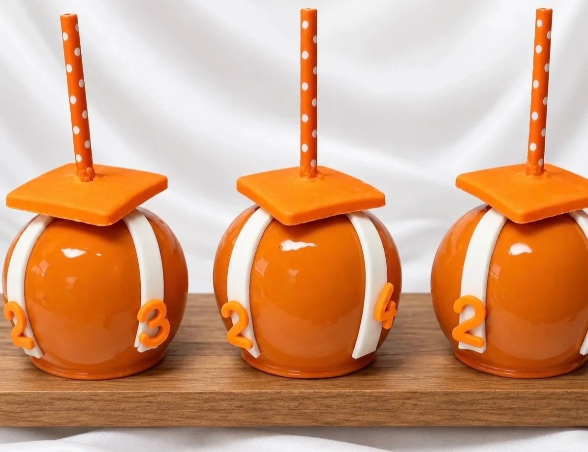 Decorative candy apples designed to resemble graduation hats with orange and white elements, each topped with a striped orange and white straw.