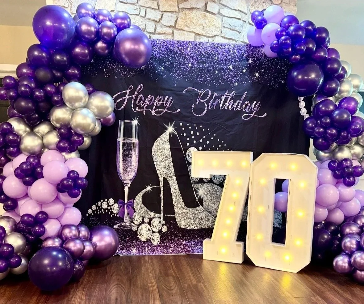 Decorative birthday backdrop with a black background, sparkles, and white text saying 'Happy Birthday.' Two large illuminated numbers, 7 and 0, represent the age 70. Balloon arrangements in shades of purple, lavender, and silver frame the backdrop.