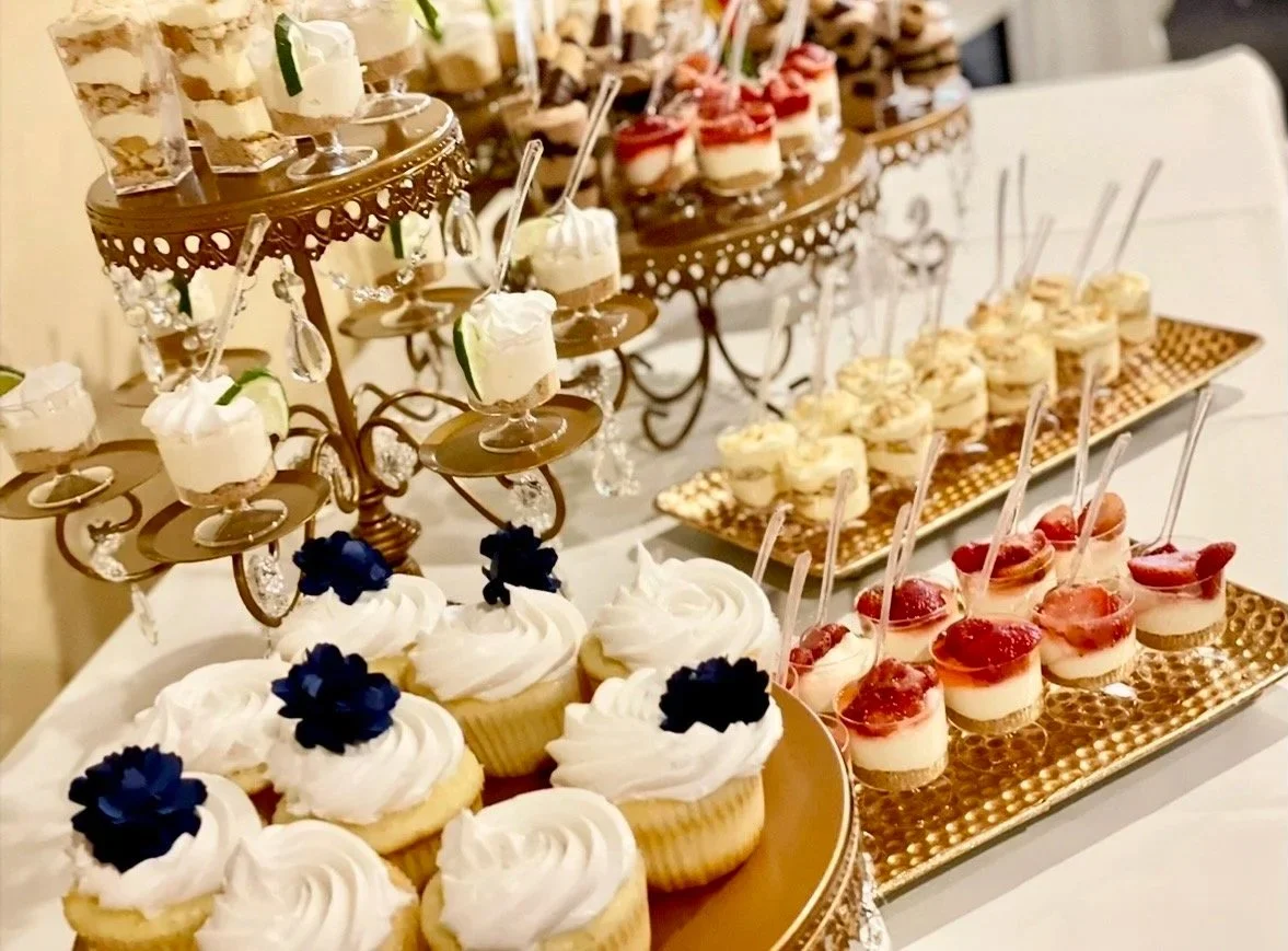 Assorted desserts on gold-trimmed serving trays, including cupcakes with white frosting and black flowers, small parfaits with white and red layers in clear cups, and mini cheesecakes topped with red fruit or syrup.