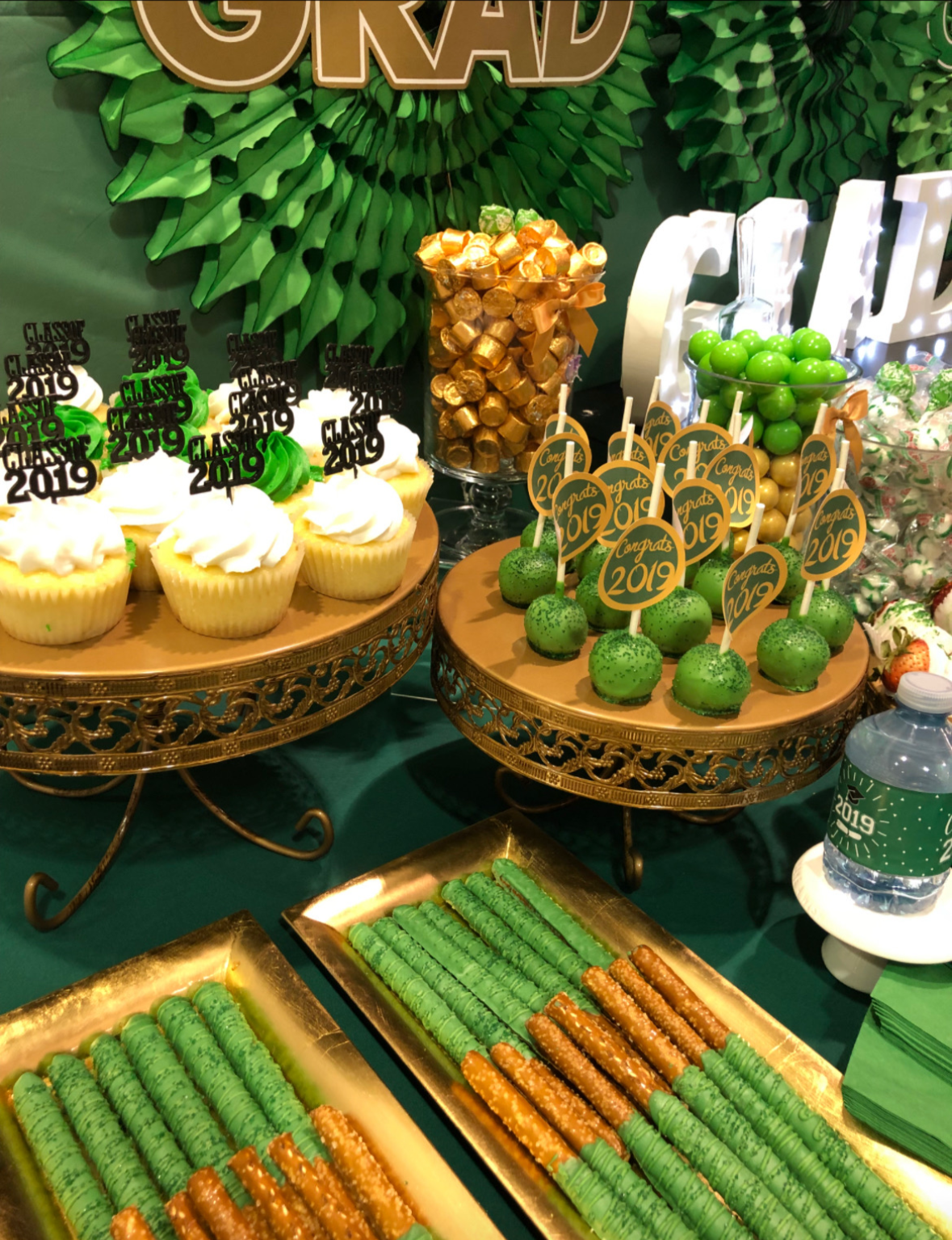 Graduation dessert table featuring cookies, cupcakes with white frosting and black toppers reading 'Class of 2019'. Green cake pops with 'Congrats 2019' toppers. Gold-wrapped candies in a jar. Green gumballs and candies with holiday wrapping.