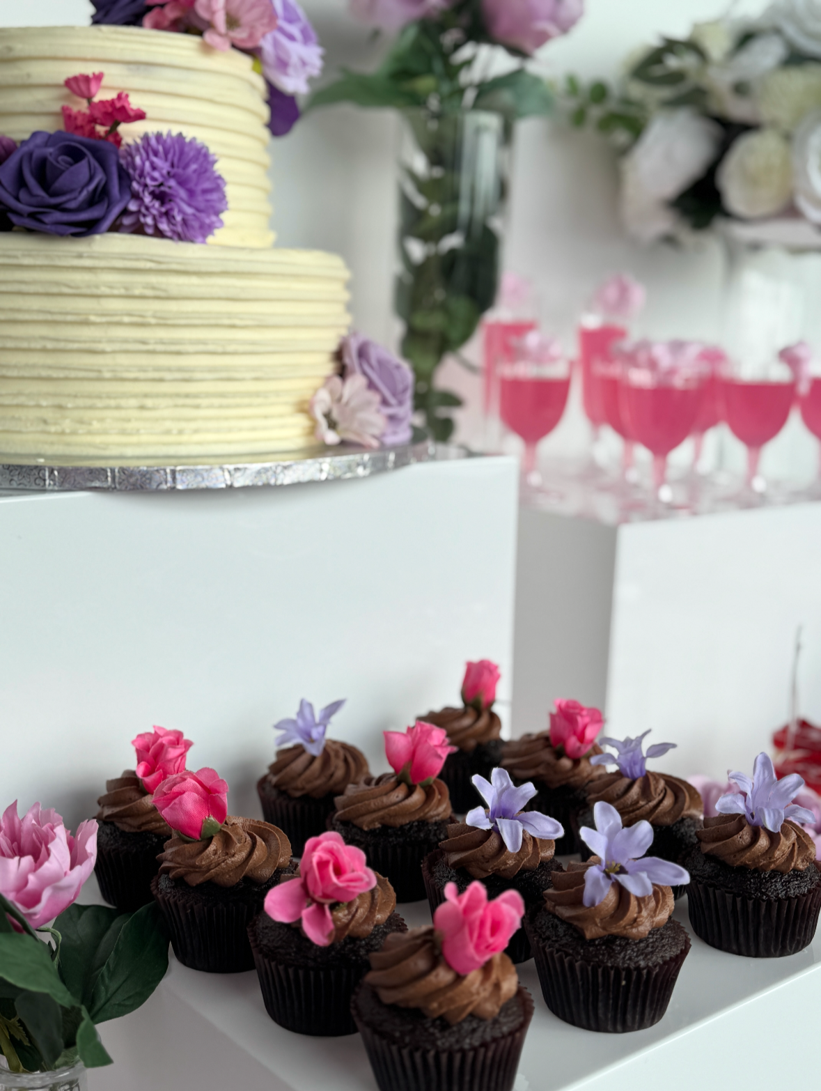 Dessert table featuring chocolate cupcakes with pink and purple flowers on top, a cake with purple and pink flowers, and glasses of pink drinks with flower garnishes, all on white display tables surrounded by floral decorations.