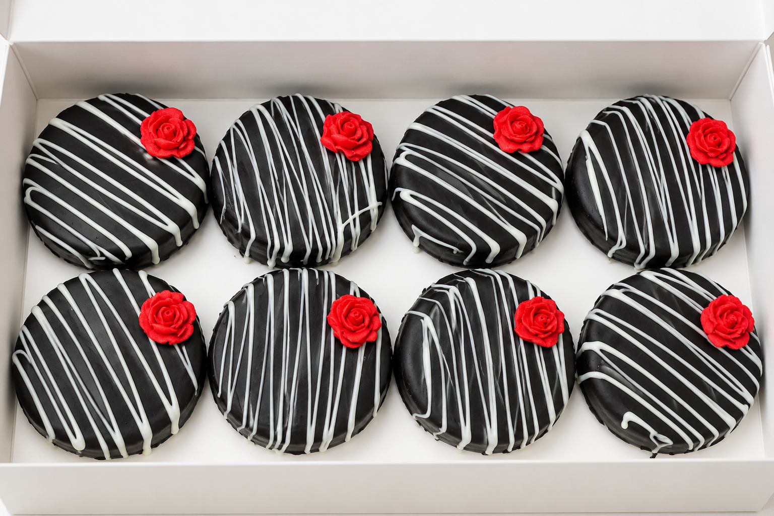 A box of eight chocolate covered oreos with black and white icing drizzle and red rose-shaped decorations.