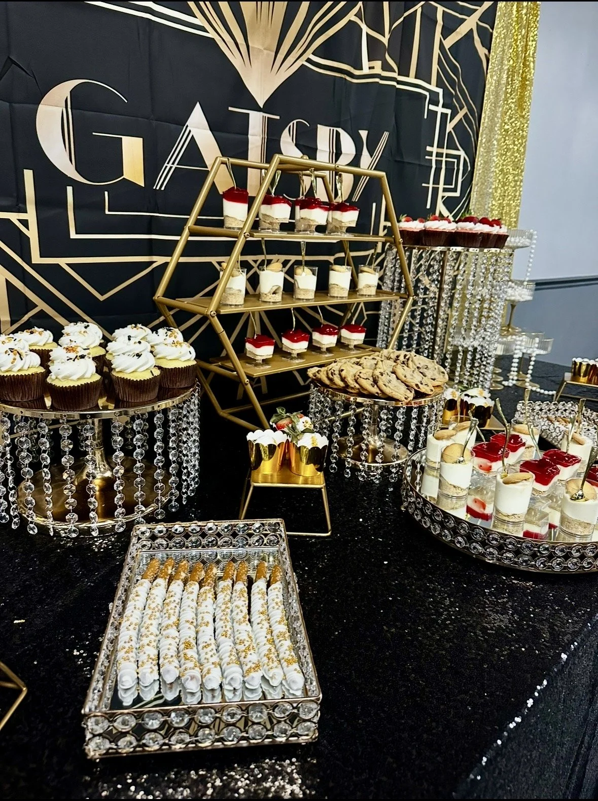Decorative dessert table with cupcakes, cookies, parfaits, and rolls, set against a black and gold geometric background with a G airy sign, featuring gold and crystal accents.
