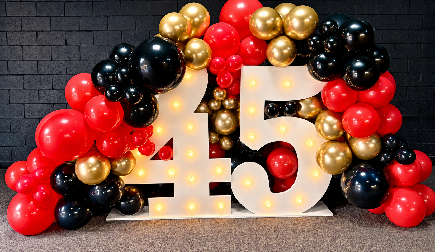 Celebratory birthday decoration with white numbers '45' illuminated, surrounded by red, black, and gold balloons against a black brick wall by Sugar Sweets by Peeks.