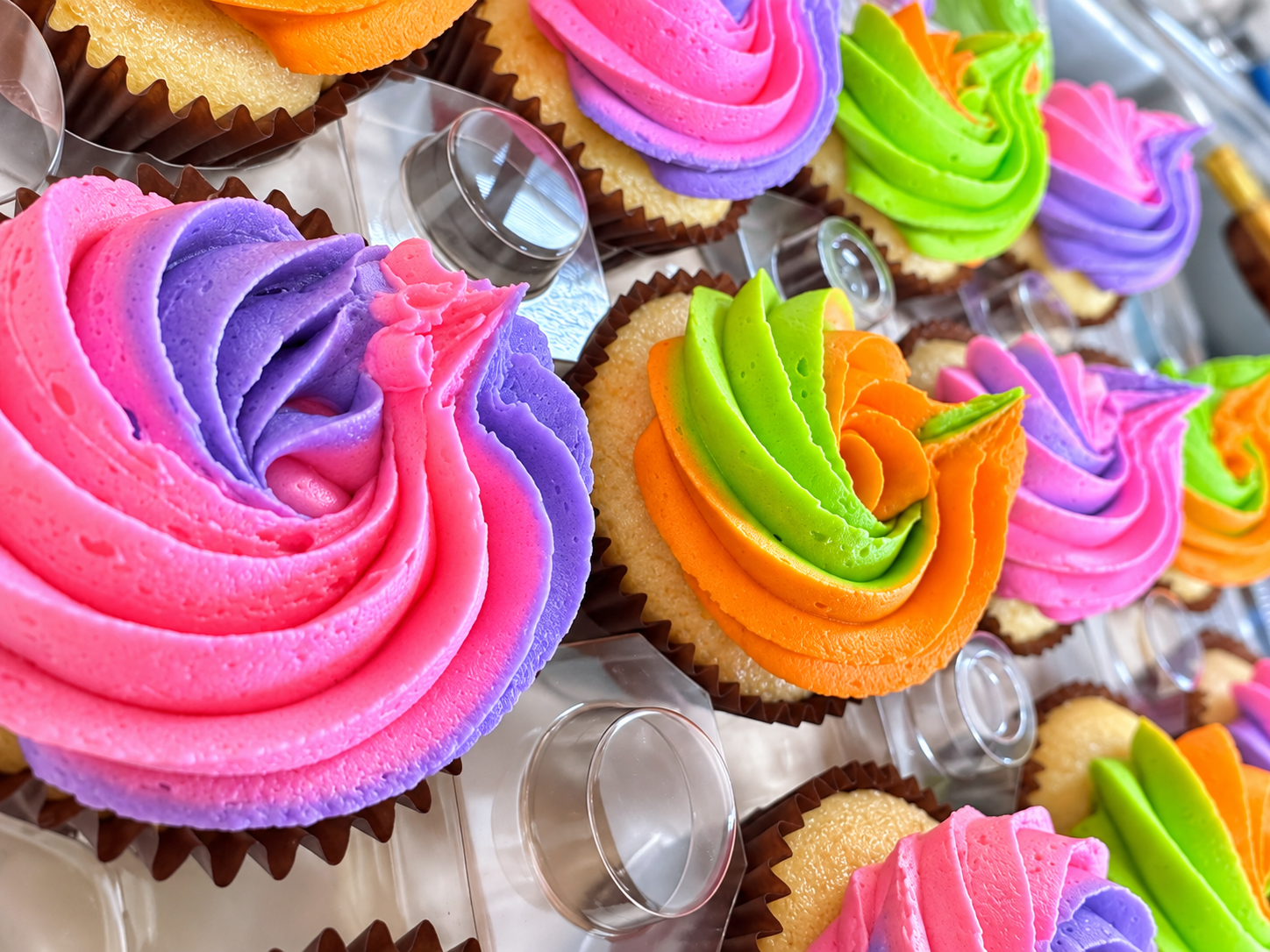Colorful cupcakes with buttercream frosting in shades of pink, purple, green, and orange, decorated in a swirl pattern, displayed on a tray.