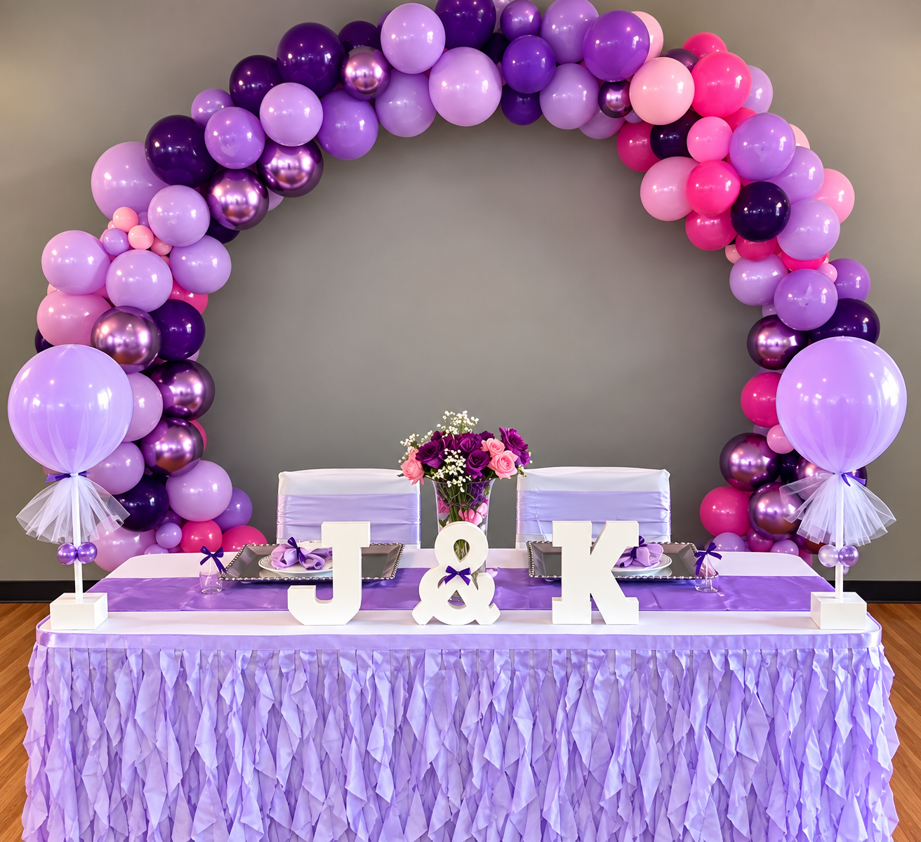 Decorative table with purple, pink, and lavender balloons arranged in an arch, featuring large letter blocks 'J' and 'K' and a vase of purple and pink flowers. The table is draped in purple ruffled fabric, set with plates, purple napkins, and lavende
