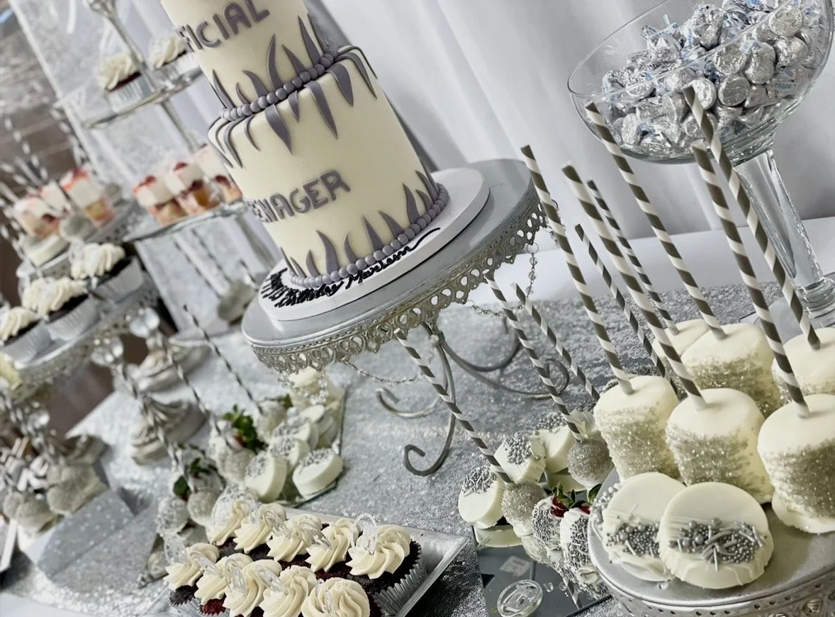 A decorated table with a large white cake with purple accents, cupcakes, and dessert treats, including chocolate truffles in a glass bowl, all in a white and silver theme for a celebration.