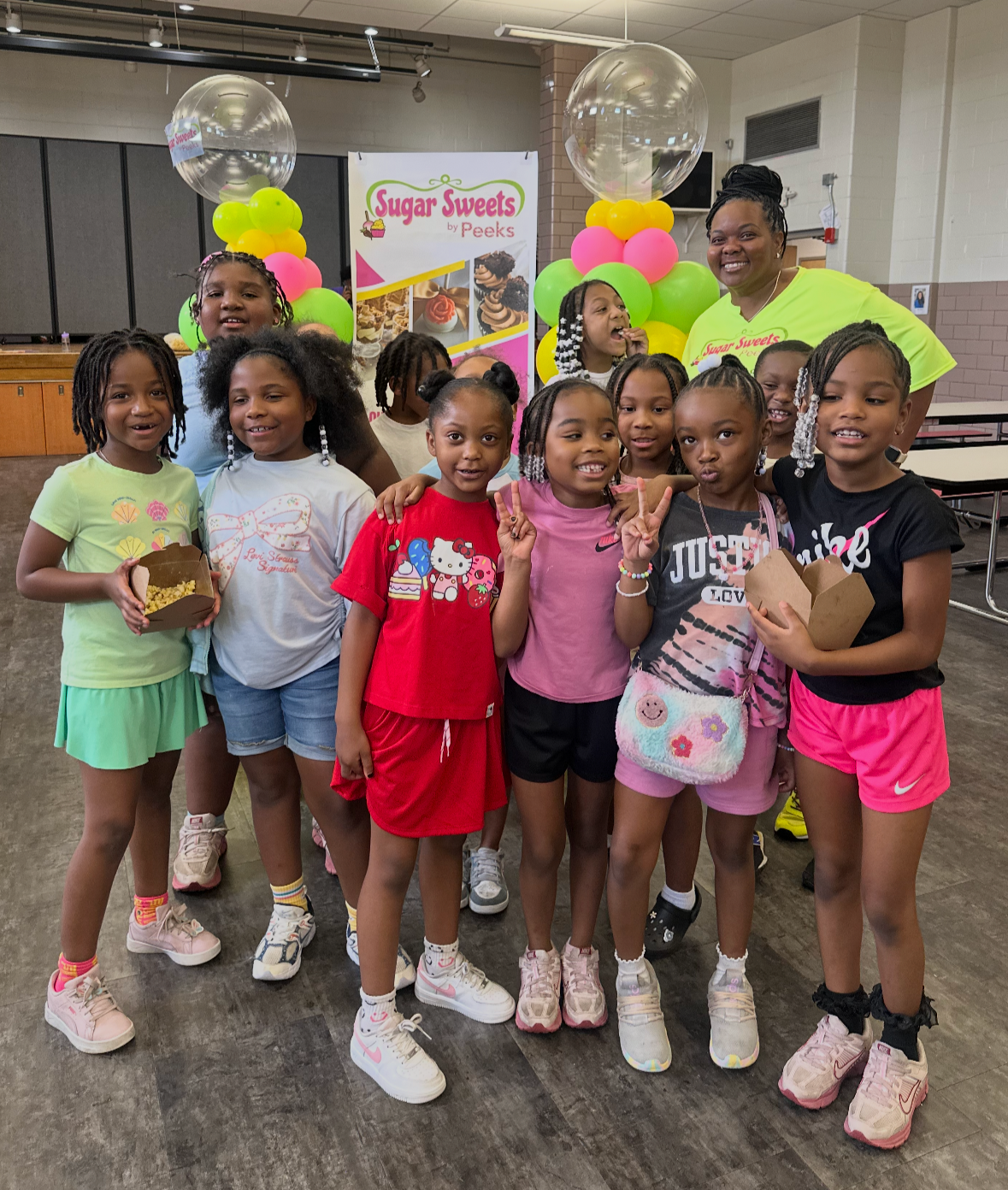 Group of class participants and Melita at a hands on Sweets class, standing in front of a colorful balloon arch and a 'Sugar Sweets' themed backdrop with cupcakes.