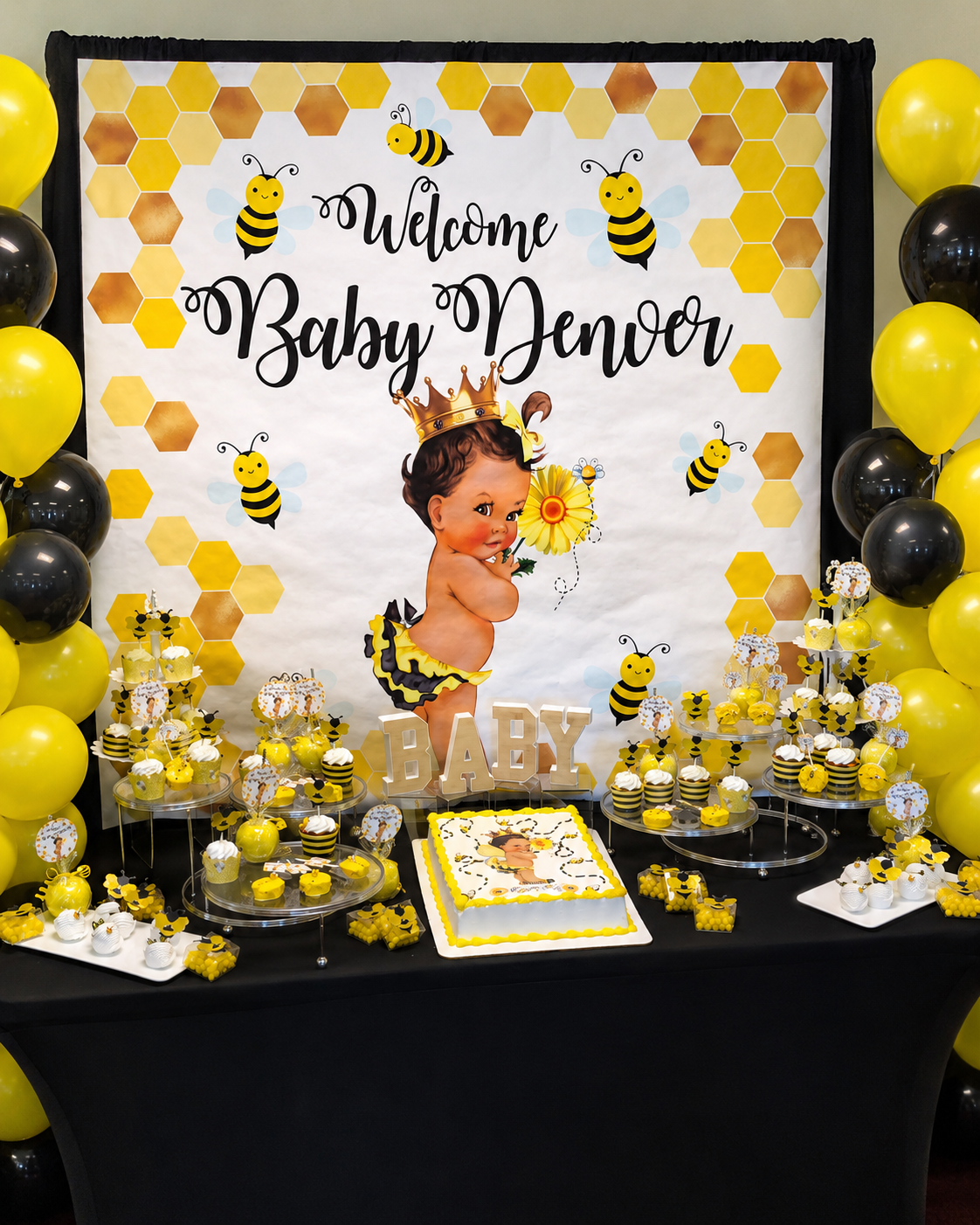 Baby shower dessert table with a honey bee theme. Features a large backdrop with a baby illustration, bees, honeycombs, and the words "Welcome Baby Denvor." The table has cupcakes, cake pops, and a decorated cake with the same bee theme. Yellow and b