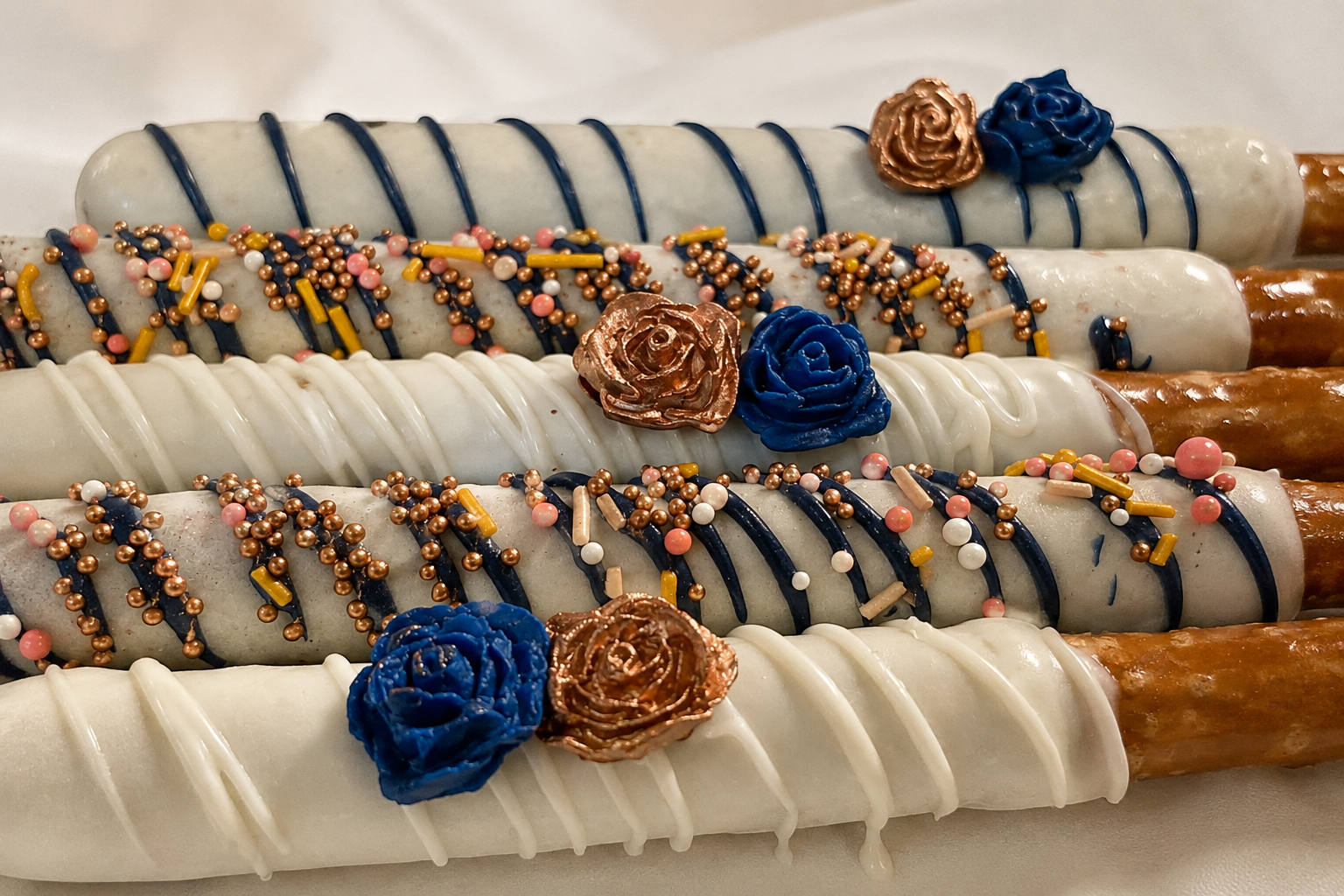 Close-up of chocolate covered pretzel rods with chocolate and caramel coatings, topped with colorful sprinkles, chocolate drizzle, and edible gold and blue roses.