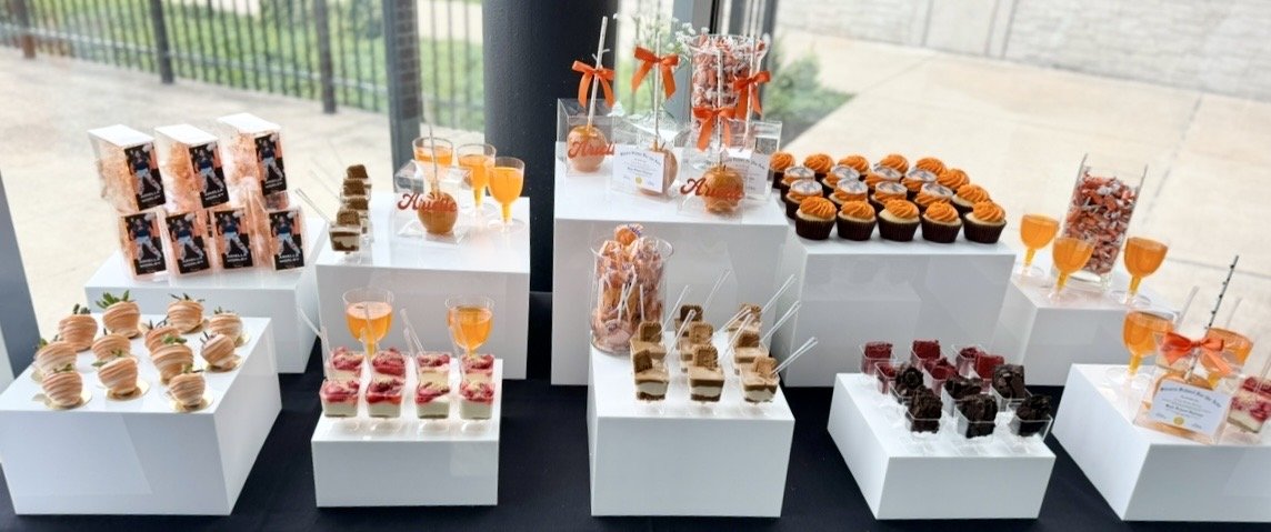 An assortment of desserts and snacks arranged on white display stands, including cupcakes, layered pastries, fruit tarts, fruit drinks, and small snack boxes, displayed in a bright setting near a large window by Sugar Sweets by Peeks. 