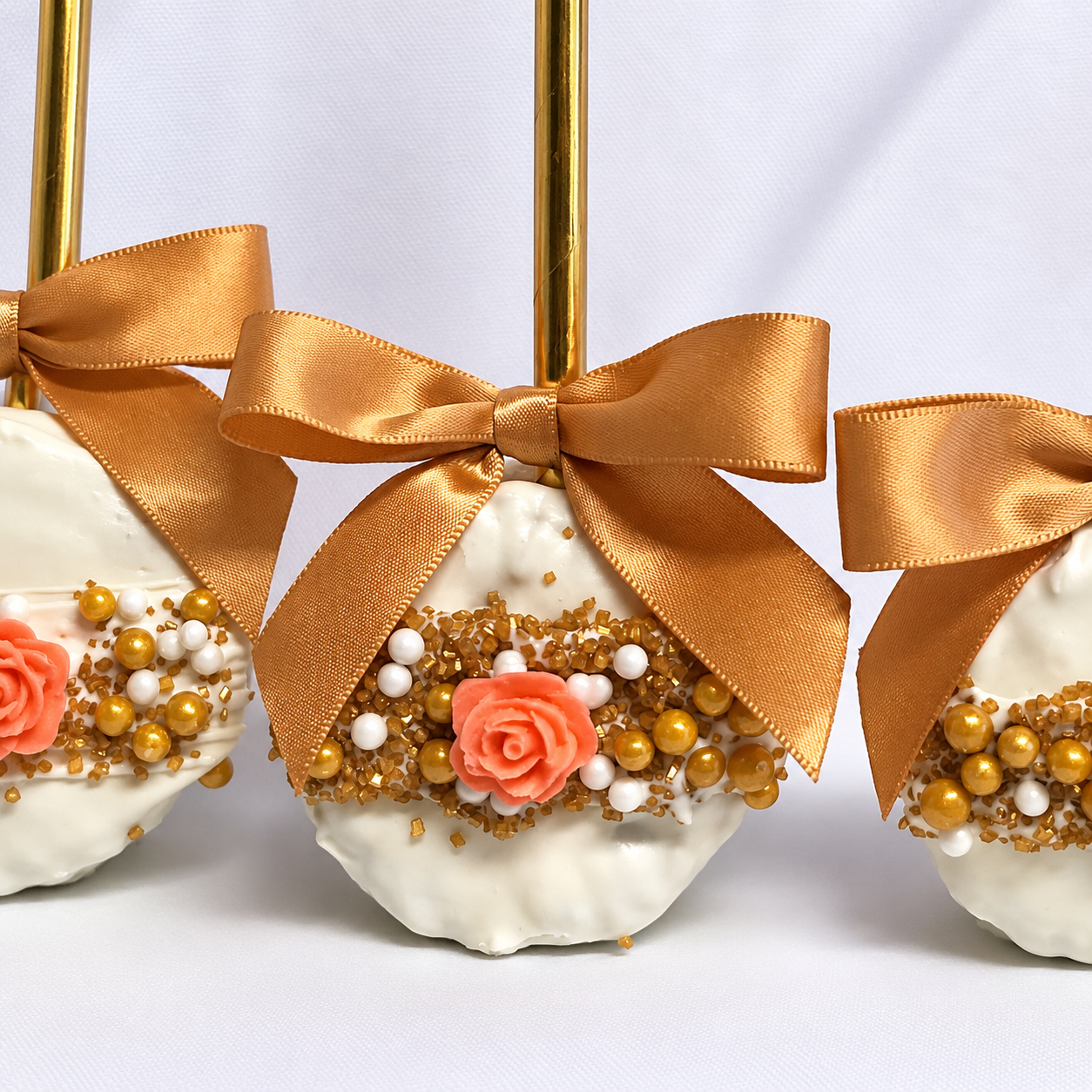 Decorative chocolate covered rice krispy treats with white coating, adorned with pink rose-shaped candies, gold and white pearls, and golden sprinkles, each topped with a gold satin bow and attached to gold sticks.